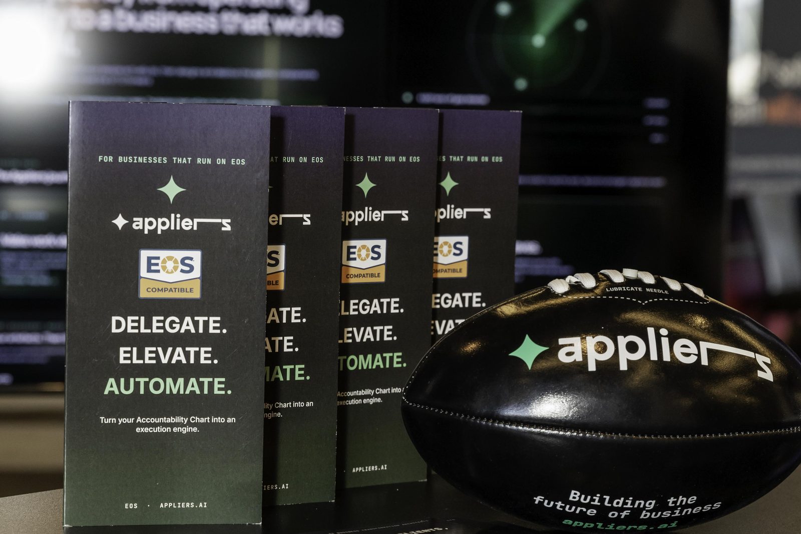 Appliers brochures fanned out on the stand table beside a branded football, printed with Delegate, Elevate, Automate.