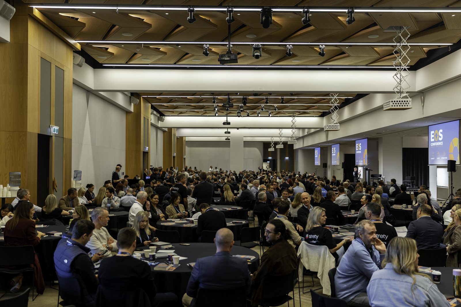 The main conference room from the back, filled with hundreds of seated attendees at round tables facing the stage.