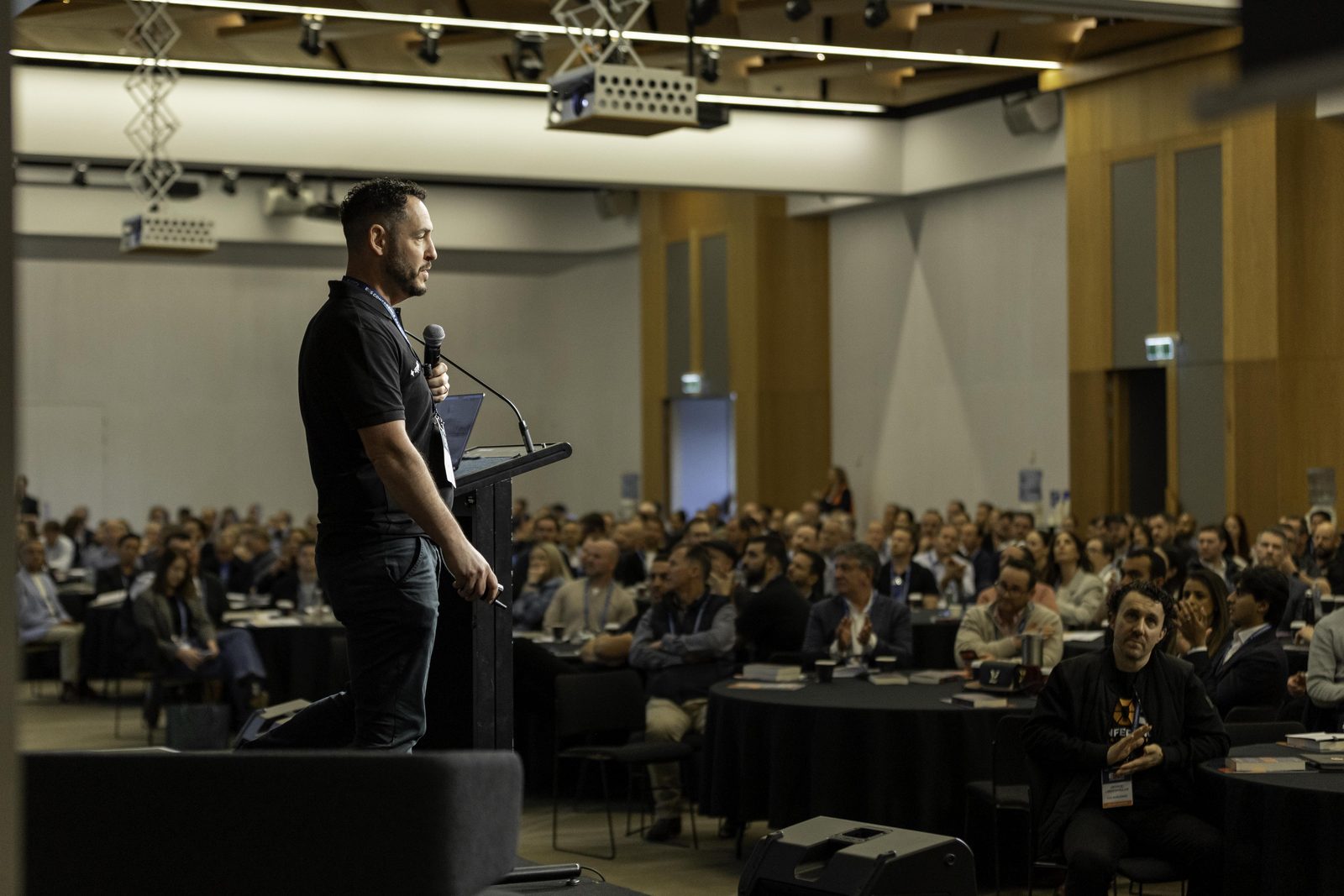 An Appliers speaker on the main stage with a microphone, addressing a full room of seated conference guests.